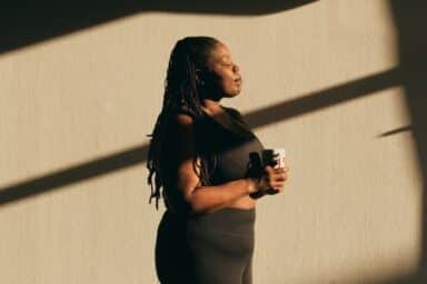 A woman stands next to a textured wall, enjoying rays of sunshine as part of her self-care routine.