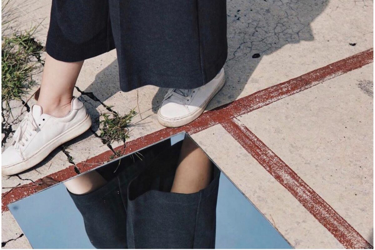 A person walks carefully next to a mirror on a sidewalk, their shoes prominently displayed, evoking a sense of covert narcissism