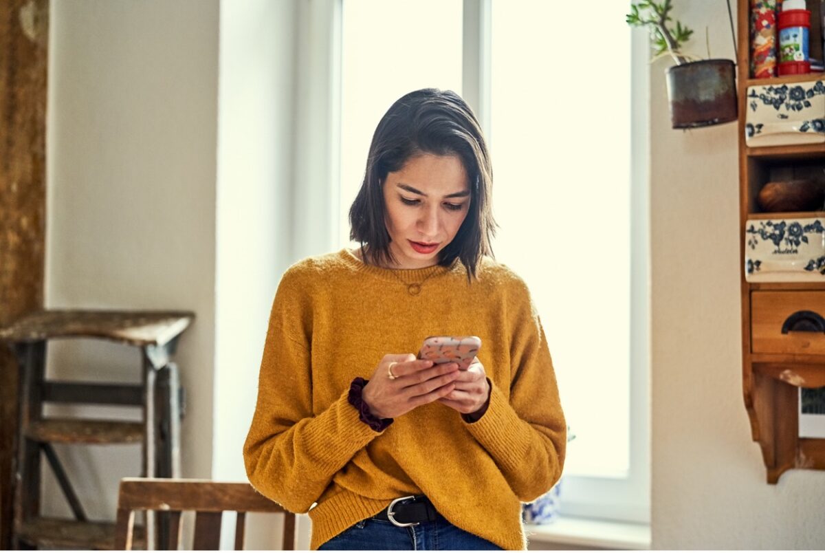 A woman standing indoors, intently focused on her phone, her troubled expression betraying her fear of missing out (FOMO)