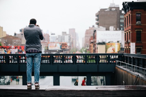 A man stands on a bridge, photographing a vibrant cityscape, suggesting a neurodivergent or different way of looking at the world.