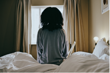 A woman is seated rigidly on the edge of a bed after a restless night, staring at a window, illustrating the link between sleep and mental health.