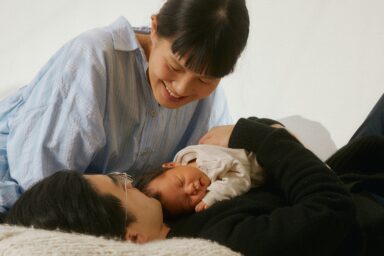 A man and woman lie on a bed, gently cradling a baby between them, showcasing a moment of family intimacy after birth