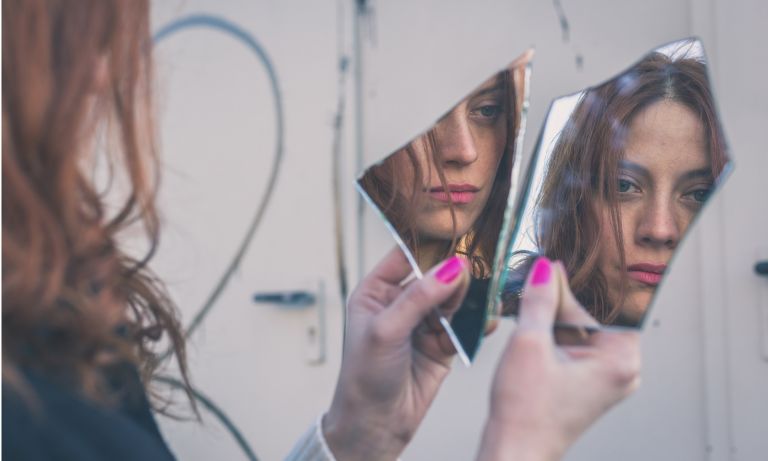 Distraught young woman holds two shards of glass in front of her, a jagged piece of her reflection visible in each