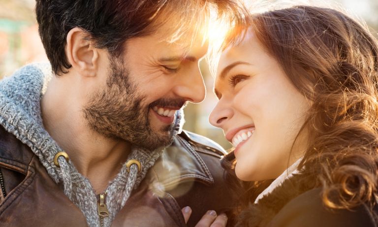 Young man and woman holding each other close and sharing smiles, her hand on his shoulder, their faces inches apart