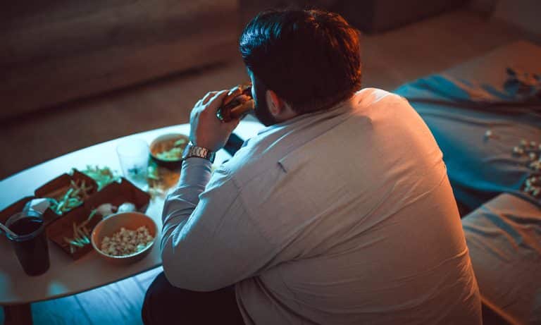 Man in dark room, eating burger with French fries, popcorn, and a glass of soda on coffee table in front of him